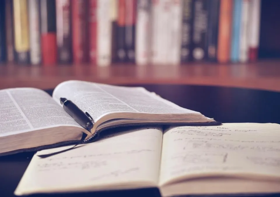 Two open books and a pen on a dark table, with a shelf of books blurred behind