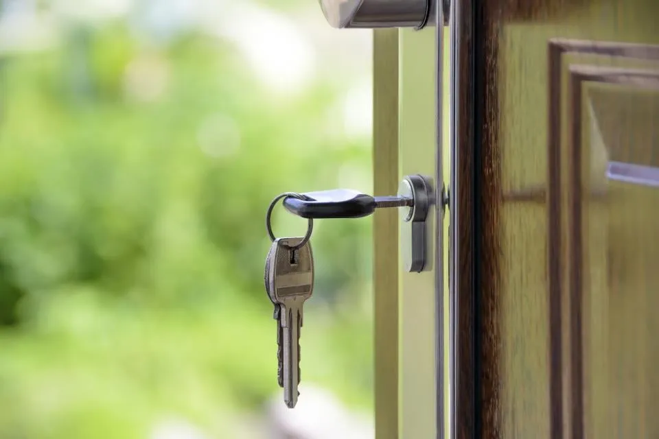 A house key hanging in the lock of an open wooden door, with green foliage blurred behind