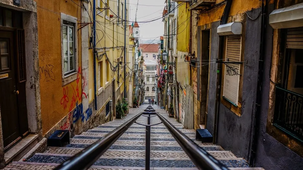 A steep cobbled lane in Lisbon between yellow and grey walls, with a handrail down the middle