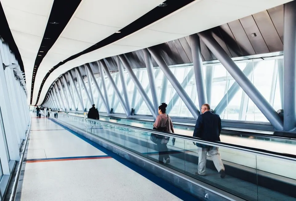 A long glass walkway in an airport with a moving walkway and a few travelers
