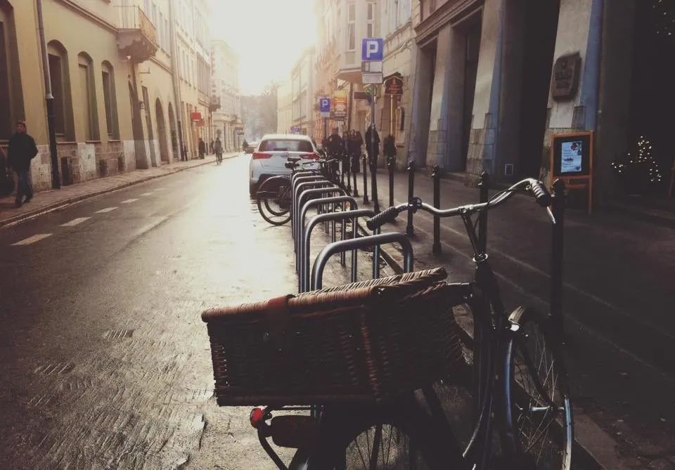 A European street in low sun with a white car parked ahead and a bicycle with a wicker basket locked to a rack