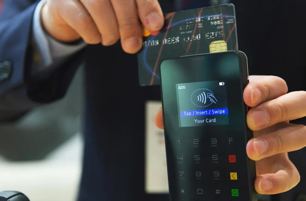 A hand holding a bank card above a handheld card terminal that prompts for a tap, insert or swipe
