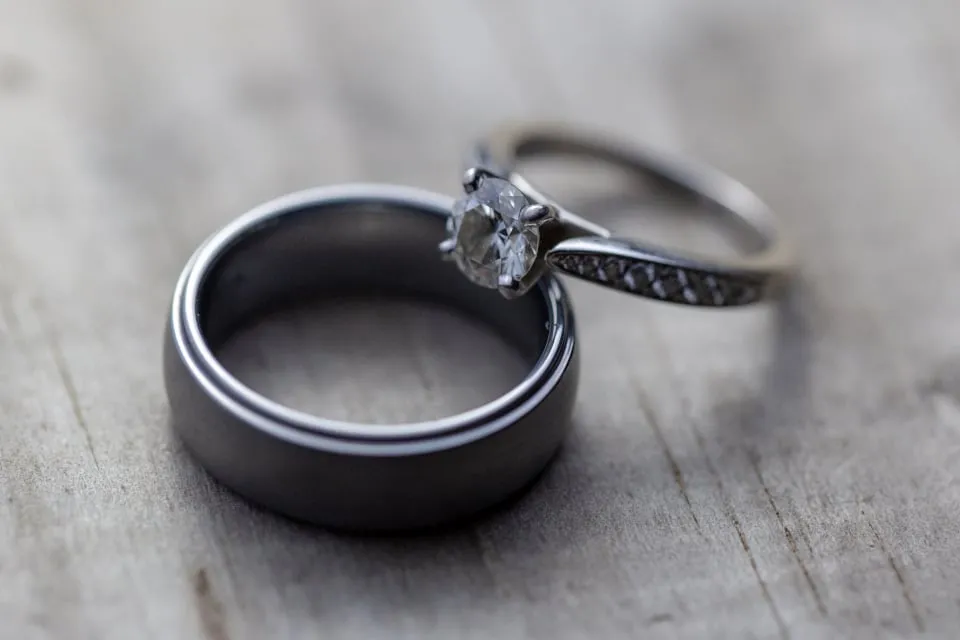 Two wedding rings, a dark band and a ring with a stone, resting on a weathered gray wooden surface