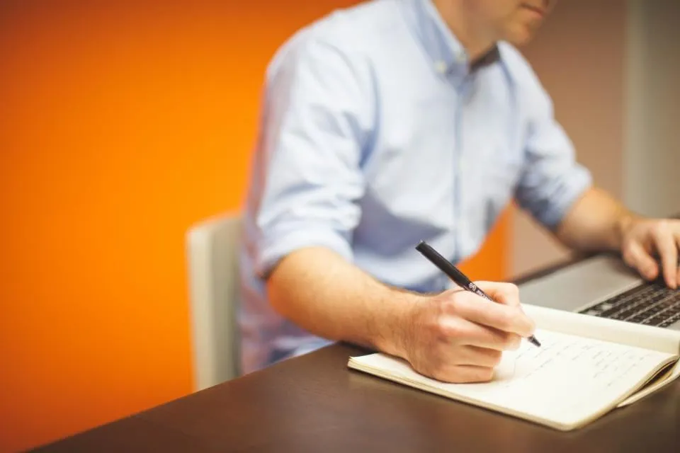 A person in a light blue shirt writing in a notebook next to an open laptop, against an orange wall