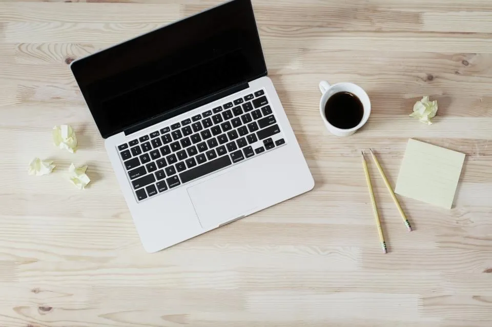 An open laptop, a cup of coffee, pencils and a notepad on a light wooden desk