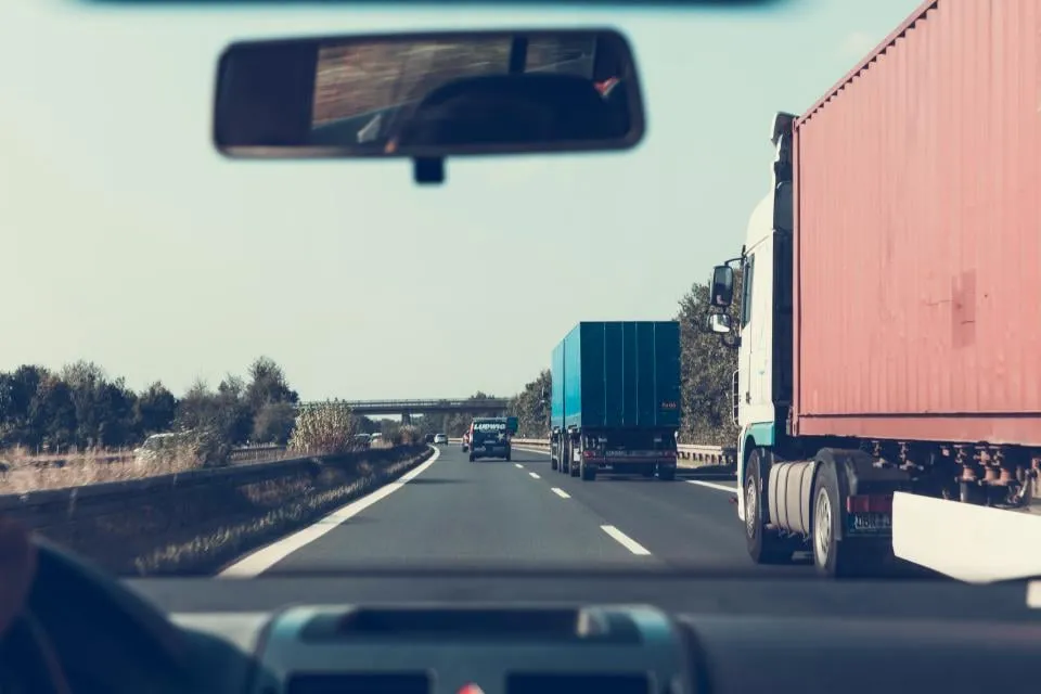 View through a car windshield of a highway with a red freight truck ahead and the rear-view mirror at the top