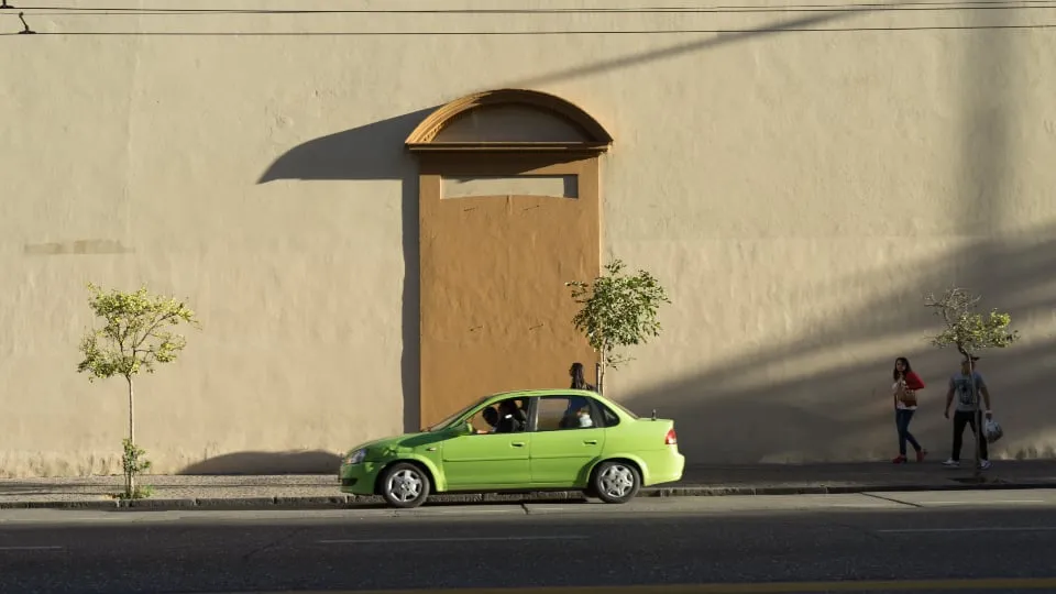 A lime green sedan parked at the curb of a long sunlit wall with two pedestrians walking past