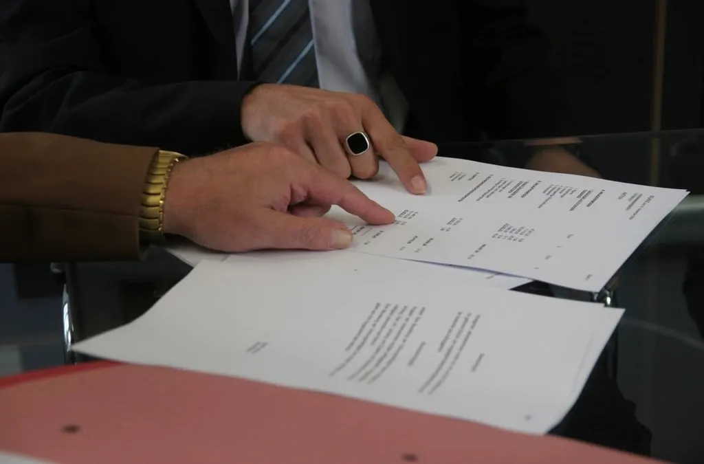 Two people at a table with printed contract pages in front of them, only their hands visible