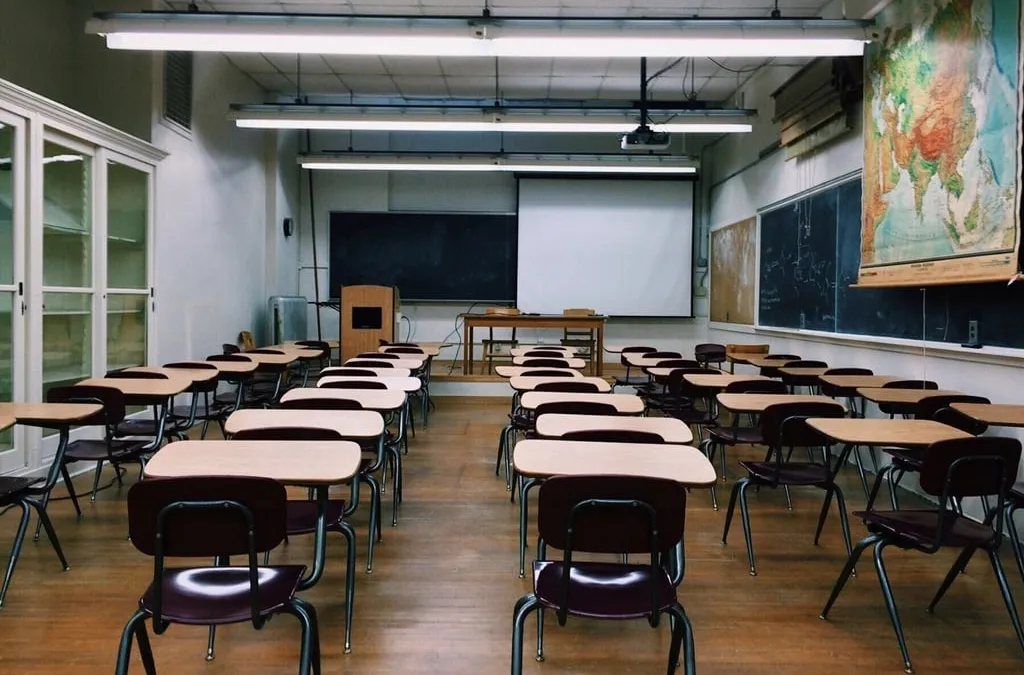 An empty classroom with rows of wooden desks and chairs, a blackboard, a projector screen and a wall map