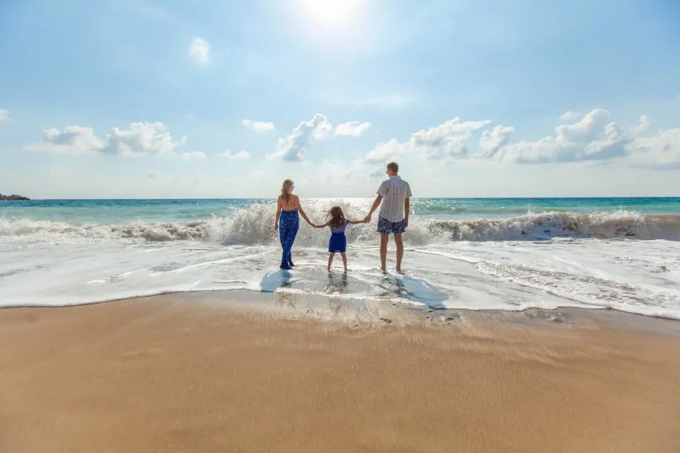 A mother, a child and a father seen from behind, holding hands as they stand in the surf on a sunny beach