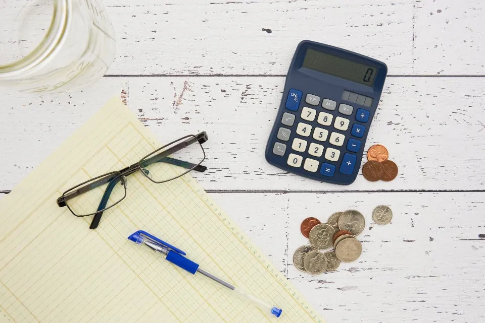 A pen, a calculator, glasses and U.S. coins on a white wooden table