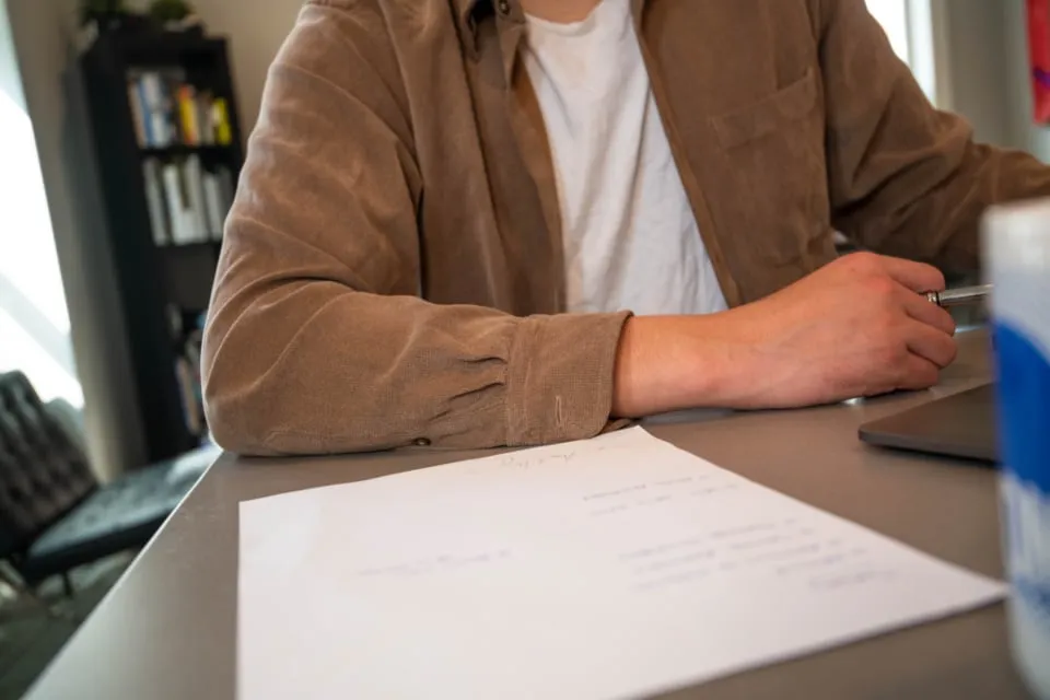 A person in a brown corduroy jacket seated at a desk with a pen in hand and a sheet of paper in front of them