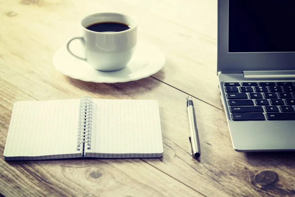 A cup of coffee, a spiral notebook with a pen and a laptop on a wooden table