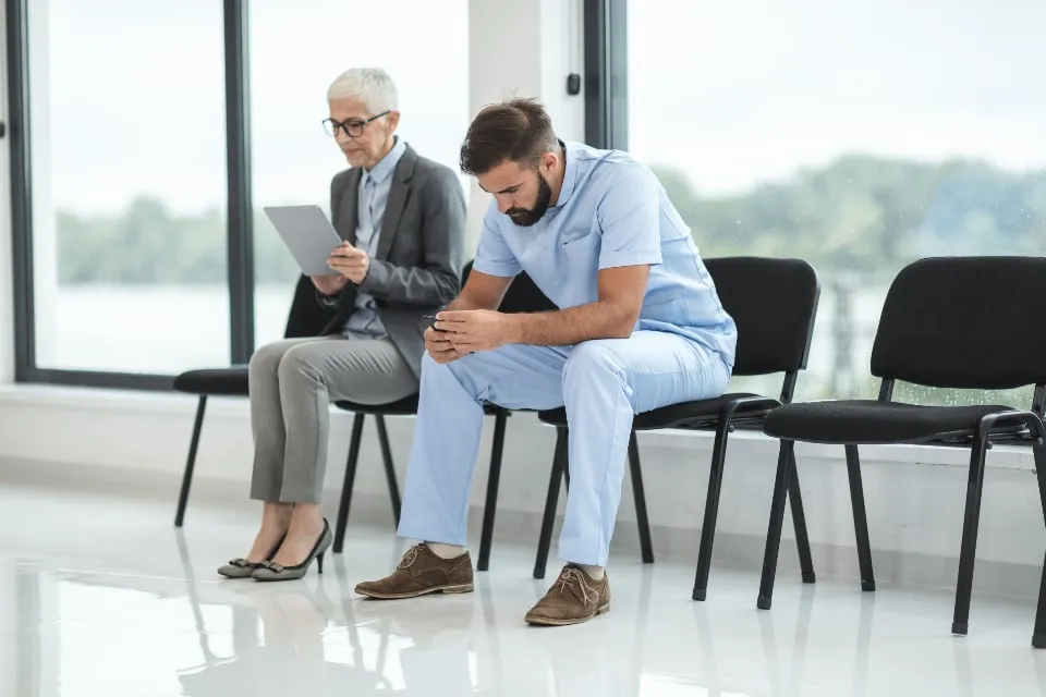 A man in scrubs and an older woman sitting on black chairs in a bright hospital waiting area