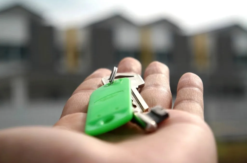An open hand holding a set of keys with a green fob in front of a blurred apartment building