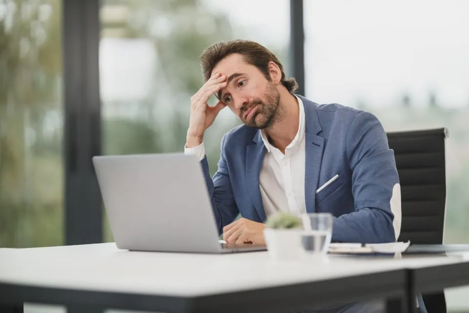 A man in a blue blazer resting his head on his hand and looking at a laptop at a white desk