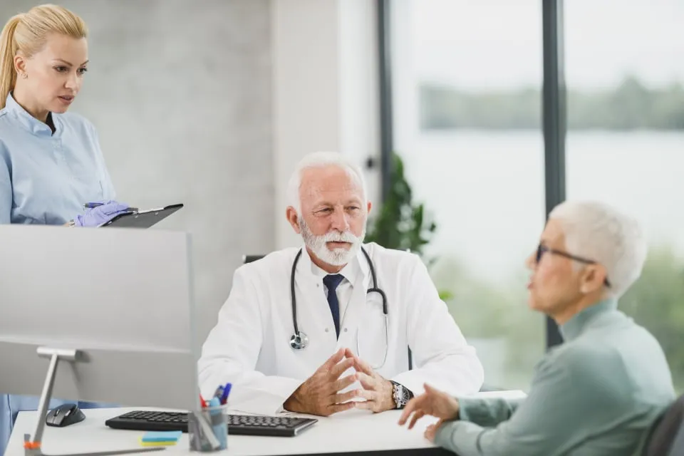 A doctor in a white coat seated at a desk talking with an older woman patient