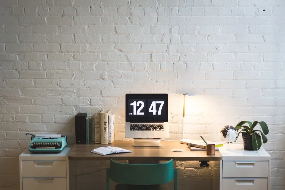 A wooden desk against a white brick wall with a laptop showing the time 12:47, a lamp, books and a teal typewriter