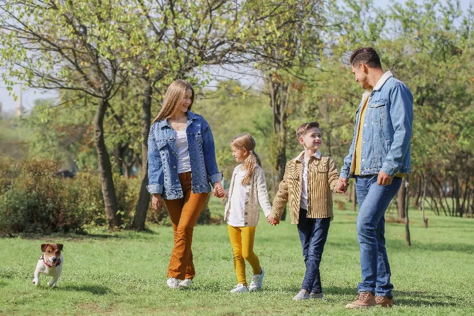 Two adults and two children holding hands and walking in a park, with a small dog