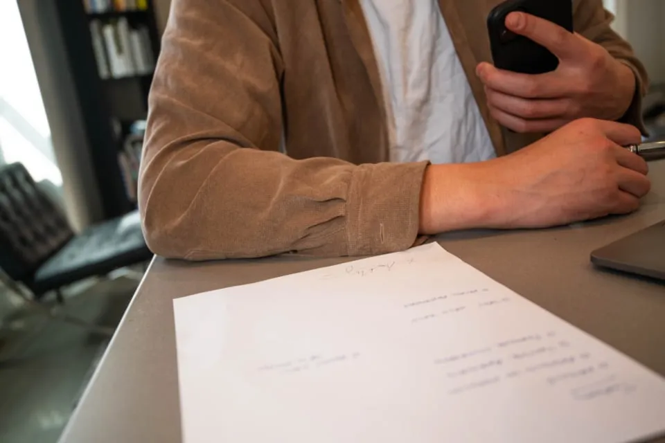 A person seated at a desk holding a pen, with a handwritten form in front of them