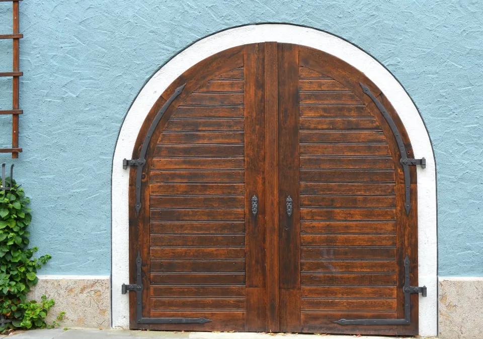 A closed arched double door of dark wooden planks with black iron hinges, set in a pale blue rough-plastered wall