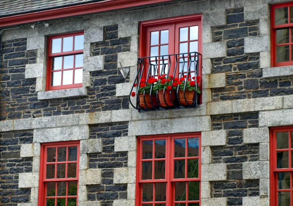 A stone building facade with red window frames and a balcony with red flowers