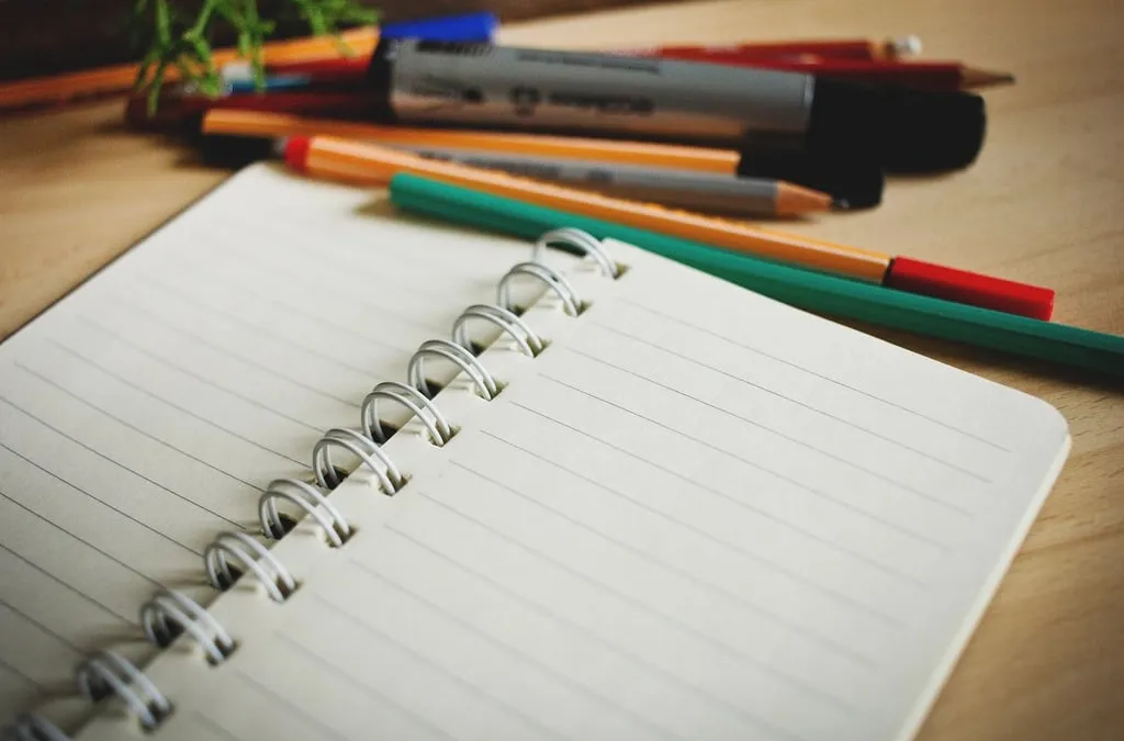 An open spiral notebook with colored pens and pencils on a wooden desk