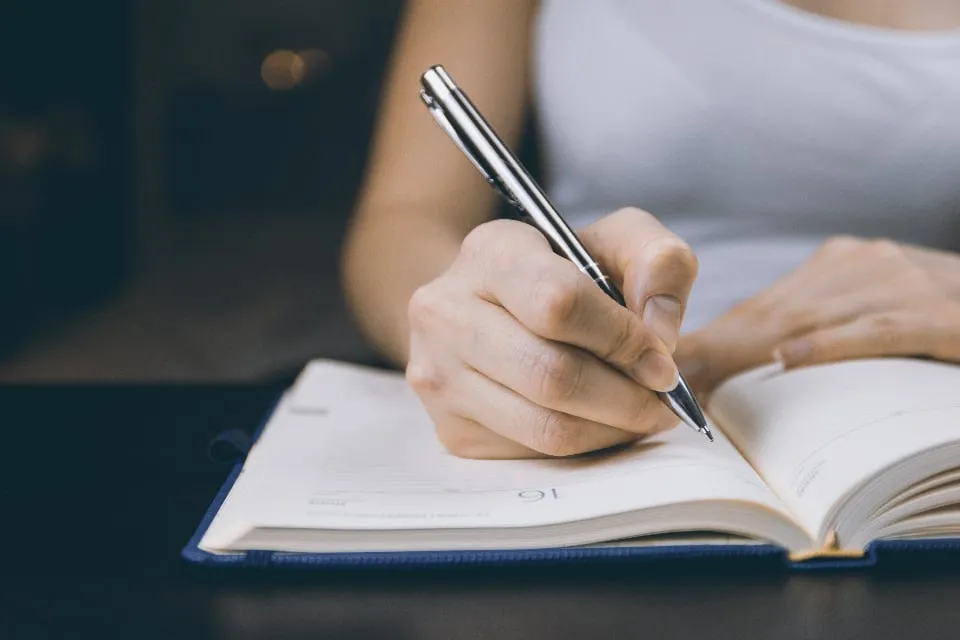 A person's hand writing with a silver pen in an open paper diary on a dark desk