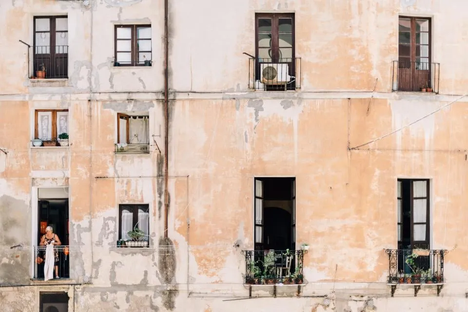 The weathered ocher facade of an apartment building with balconies and a woman at an open window