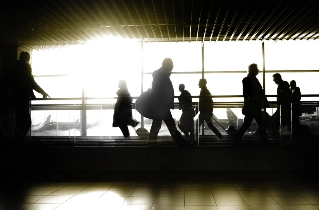 Silhouettes of travelers walking past an airport window against bright light