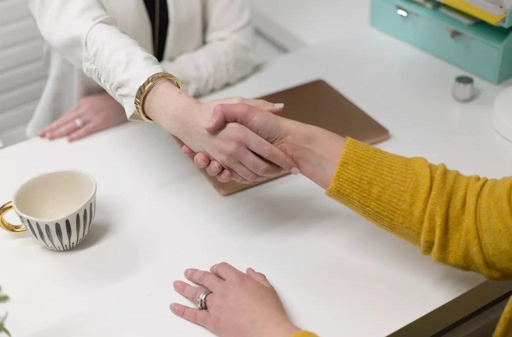 Two people shaking hands across a white table in an office