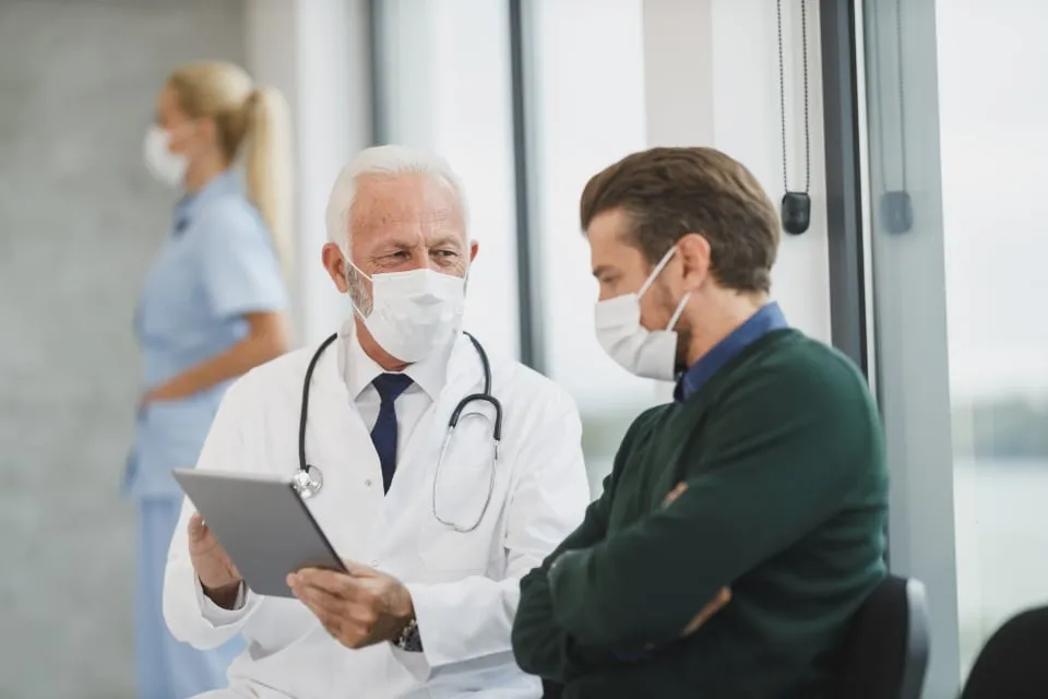 An older doctor in a white coat showing a tablet to a masked patient in a bright clinic