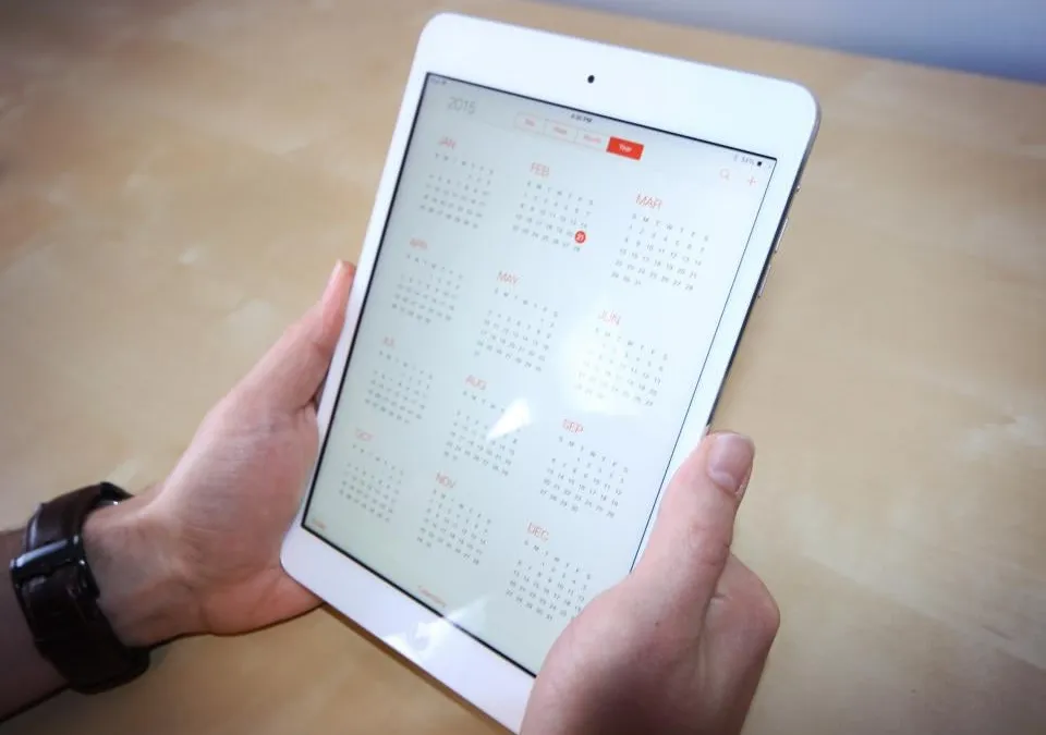 Two hands holding a white tablet that shows a year calendar, January to December, on a light wooden table