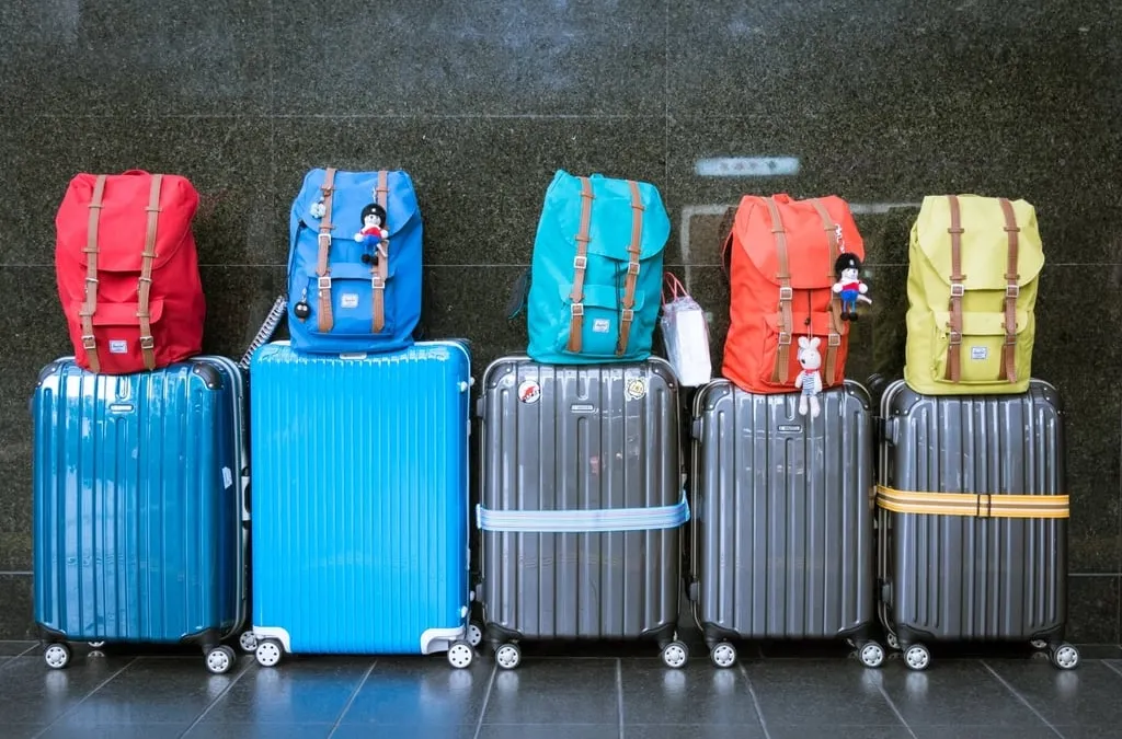 Five rolling suitcases in blue and gray with colorful backpacks on top, lined up against a dark wall