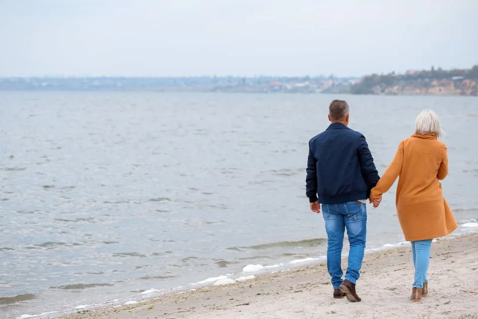 An older couple walking hand in hand on a beach, seen from behind
