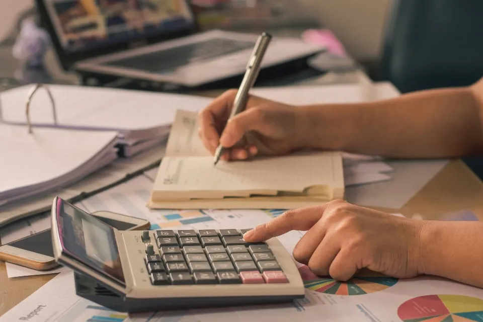 Hands writing in a notebook while using a desktop calculator, with papers and a laptop on the desk