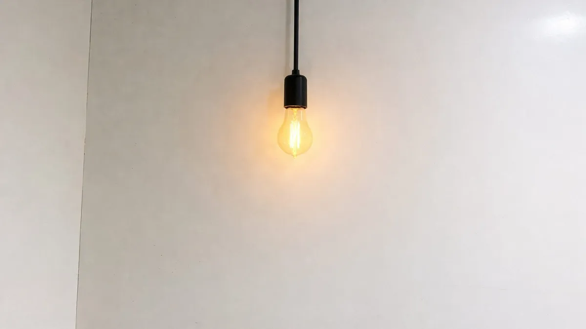 A single glowing filament light bulb hanging from a black cord against a plain white wall