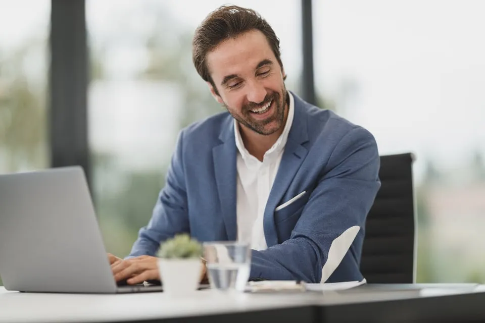 A smiling man in a blue blazer typing on a laptop at a white office desk
