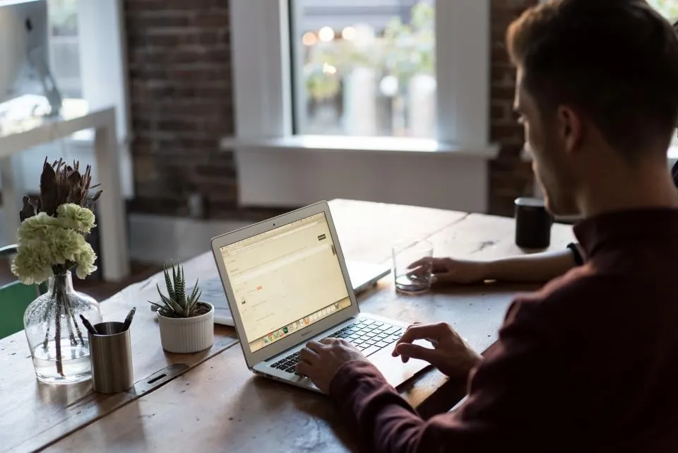 A man typing on a laptop at a wooden table by a window